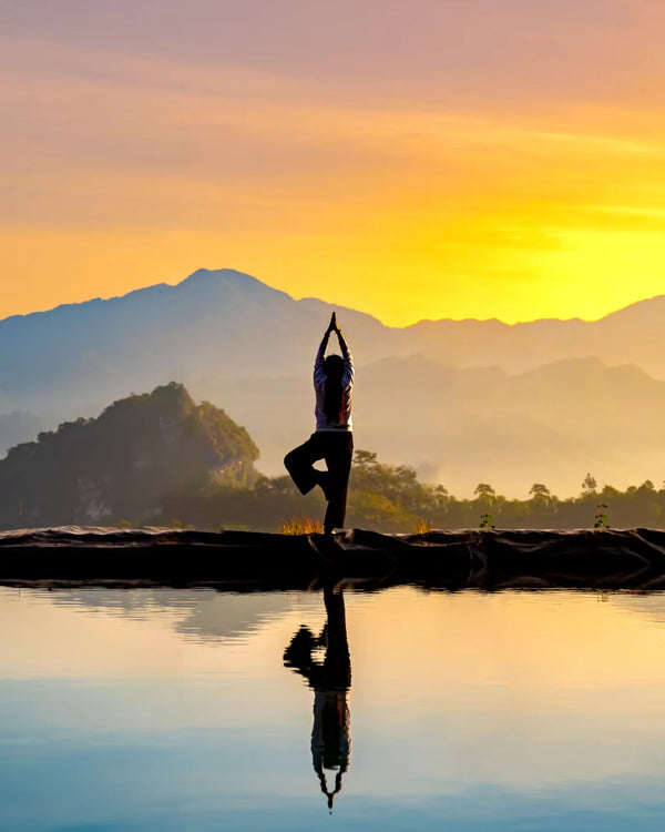 A person practicing yoga at sunrise on a calm, reflective surface, with mountains in the background